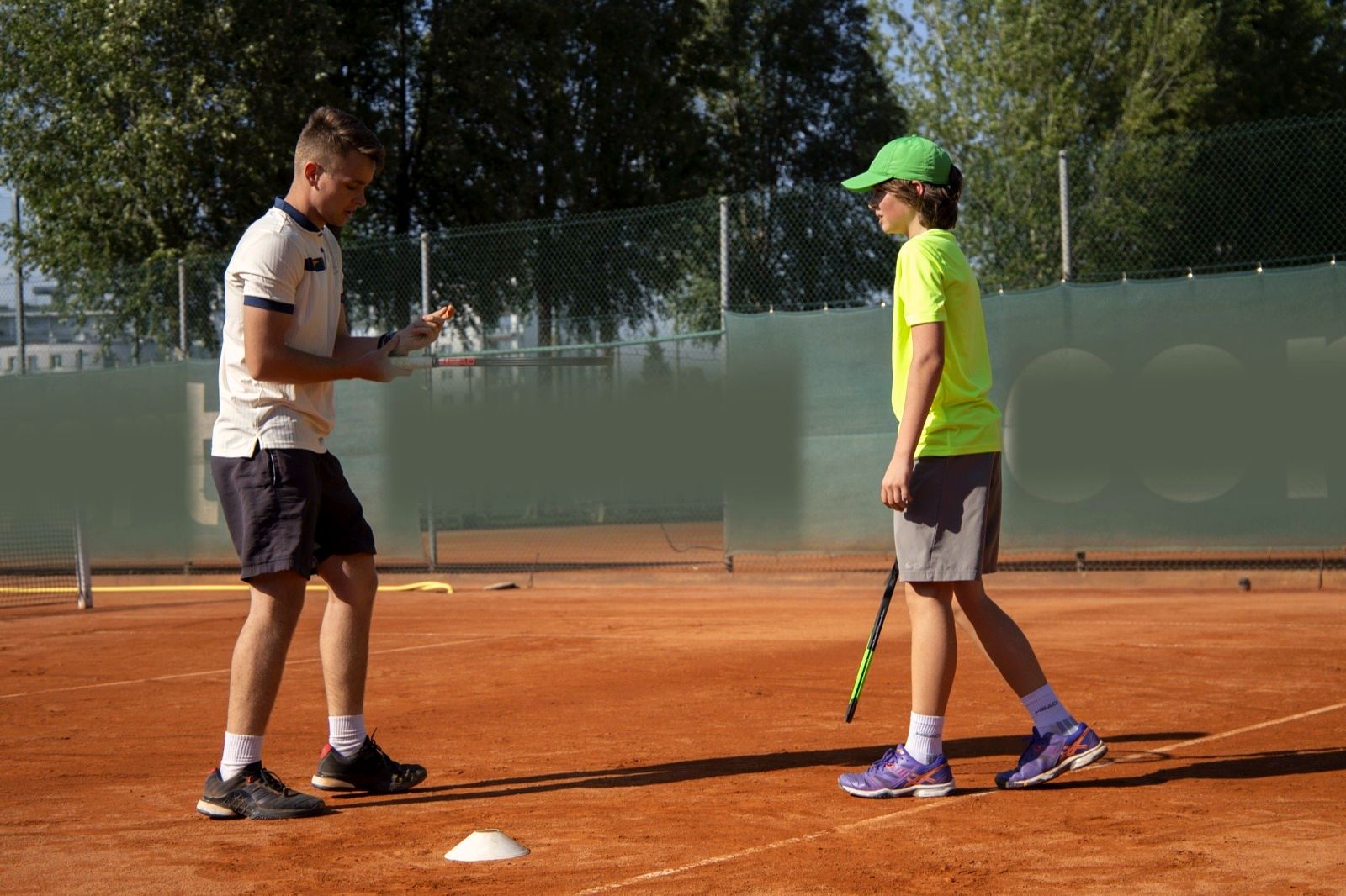 Ein Coach erklärt einem Jugendlichen mit grüner Kappe eine Übung auf dem Sandplatz